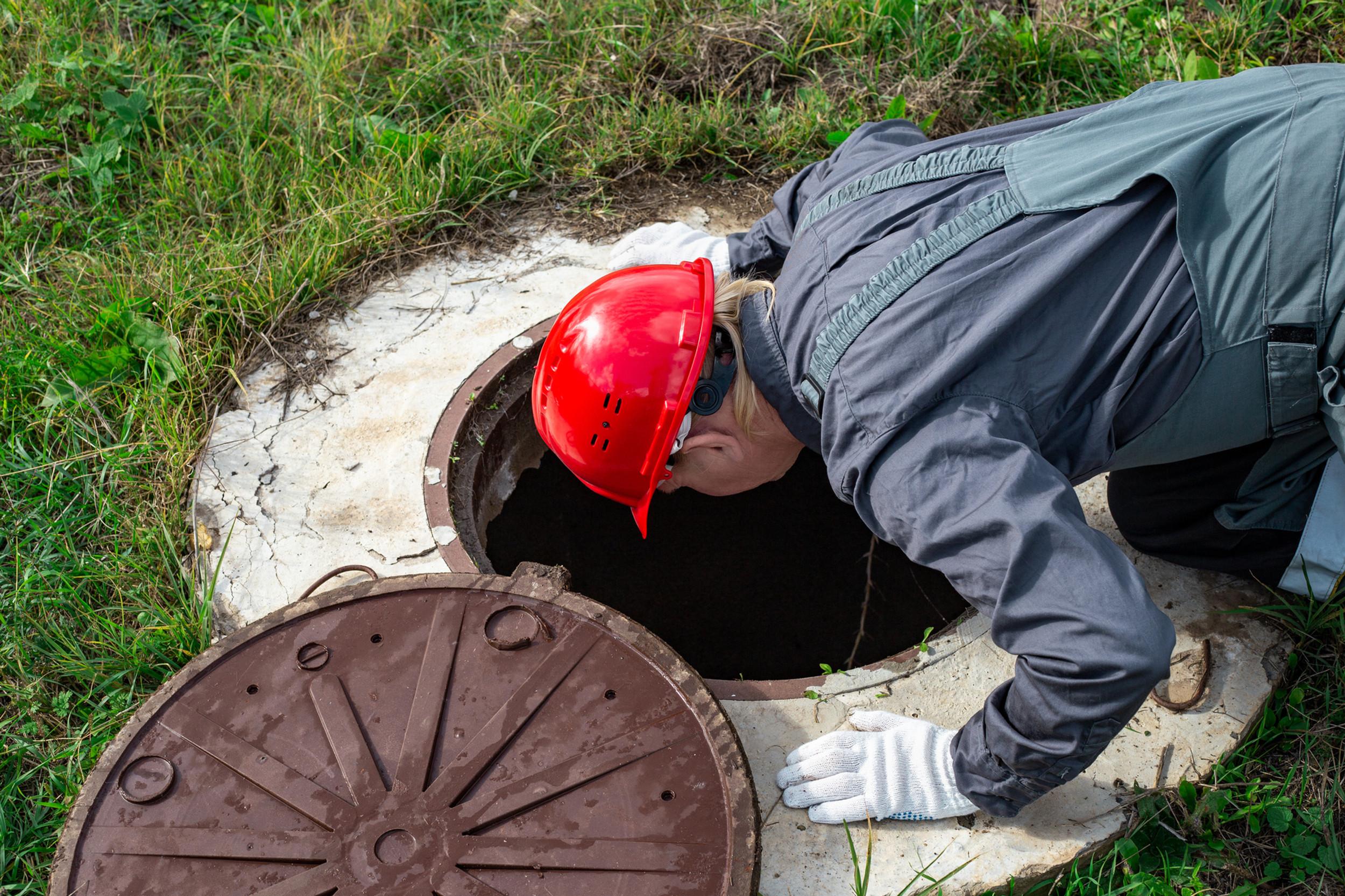 Anytime Septic technician in a red hard hat inspecting an open septic tank access lid in the yard
